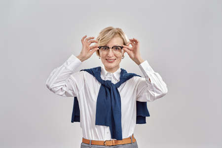 Portrait of elegant middle aged caucasian woman wearing business attire touching her glasses, smiling at camera while standing isolated over grey backgroundの写真素材