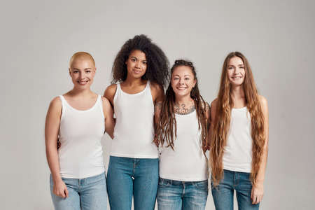 Portrait of four young diverse women wearing white shirts and denim jeans smiling at camera while posing together isolated over grey backgroundの写真素材