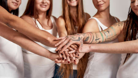 Cropped shot of diverse women in white shirts showing unity, putting hands on top of each other like a team while standing isolated over grey backgroundの写真素材