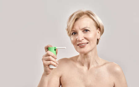 Portrait of attractive middle aged woman smiling at camera and holding throat spray, standing isolated over grey backgroundの写真素材