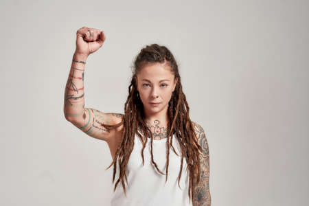 Portrait of young tattooed caucasian woman with dreadlocks wearing white shirt, looking confident at...