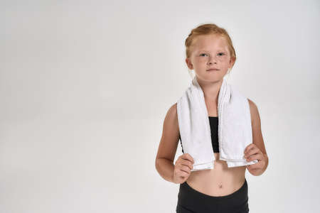 Pretty little red haired sportive girl in sportswear looking at camera, standing with white towel around her neck while posing isolated over white backgroundの写真素材