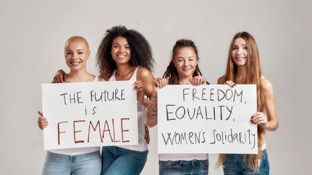 Four happy diverse women smiling at camera while holding, standing with banners in their hands isolated over grey background. Feminism and equality conceptの写真素材