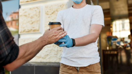 Close up of hands of man receiving hot coffee drink from cafe assistant in protective gloves while collecting his takeaway order during coronavirus lockdownの写真素材