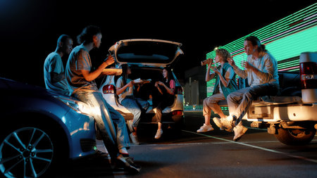 Young casually dressed friends of different nationalities sitting in opened car trunks in front of...