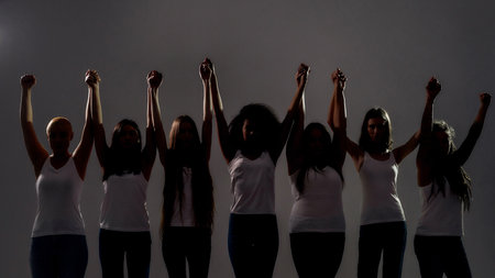 Silhouetted group of diverse women holding, raising their arms together while standing over grey background. Diversity, unity conceptの写真素材