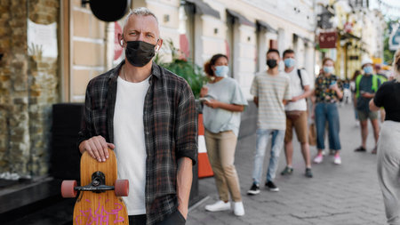 Portrait of middle aged man wearing mask holding longboard, looking at camera while waiting to collect his takeaway order from the pickup point during lockdownの写真素材