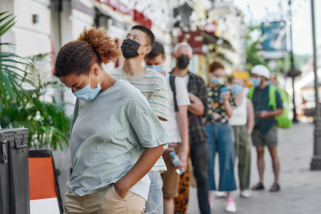 Young african american woman wearing mask waiting, standing in line with other people, respecting social distancing to collect takeaway order from the pickup point during lockdownの写真素材