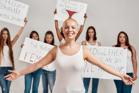 Young caucasian woman with shaved head in white shirt looking excited. Group of diverse women holding protest banners for woman power and rights in the backgroundの写真素材
