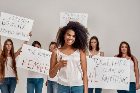 Young african american woman in white shirt showing OK sign, smiling at camera. Group of diverse...