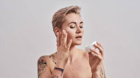 Soft Touch. Portrait of beautiful tattooed woman with pierced nose and short hair applying skin care product on face, holding white plastic jar of cream isolated over grey backgroundの写真素材