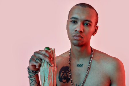 Drink responsibly. A dark-skinned man with tattoos holding a bottle in a paper bag looking into a...