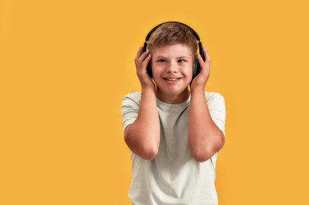 Happy disabled boy with Down syndrome in headphones listening to music and smiling aside while posing isolated over yellow backgroundの写真素材
