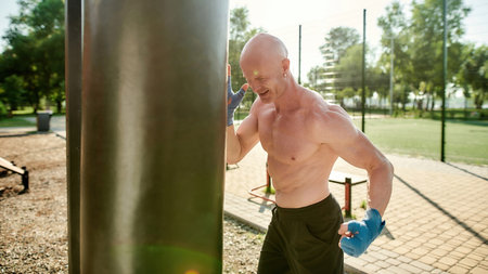 Focused mature man boxing with black punching bag, having workout at street gym yard