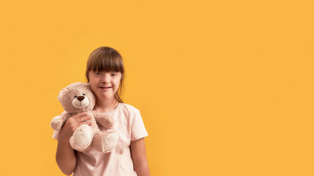 Portrait of disabled girl with Down syndrome smiling at camera, holding her teddy bear while posing isolated over yellow backgroundの写真素材