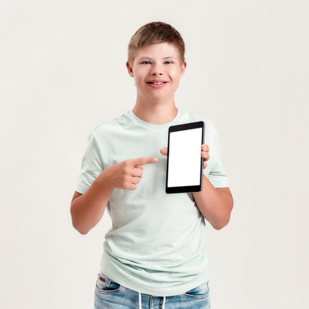 Happy disabled boy with Down syndrome smiling, holding and pointing at tablet pc with blank screen while standing isolated over white backgroundの写真素材