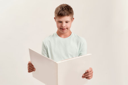 Teenaged disabled boy with Down syndrome looking focused while reading a book, standing isolated over white backgroundの写真素材