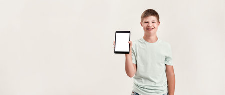 Happy disabled boy with Down syndrome smiling, holding and showing tablet pc with blank screen while standing isolated over white backgroundの写真素材