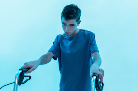 Portrait of teenaged disabled boy with cerebral palsy looking at camera, taking steps with his walker isolated over blue background with neon lightの写真素材