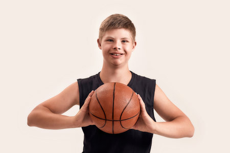 Cheerful disabled boy with Down syndrome smiling at camera, holding basketball while posing isolated...