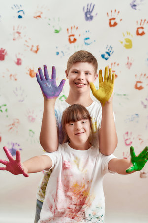 Happy kids, disabled boy and girl with Down syndrome smiling at camera, showing their hands painted in colorful paints for hand prints on the wallの写真素材