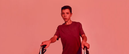 Cropped shot of teenaged disabled boy with cerebral palsy looking away, taking steps with his walker isolated over red light backgroundの写真素材