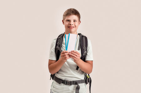 Happy disabled boy with Down syndrome wearing backpack smiling while holding air tickets, standing isolated over white backgroundの写真素材