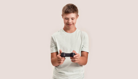 Happy disabled boy with Down syndrome smiling while holding, using game remote control, playing video games, standing isolated over white backgroundの写真素材