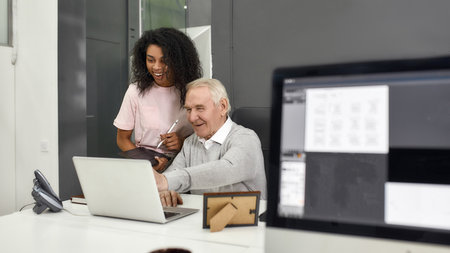 Aged man, senior intern looking at laptop while showing results to his young colleague, Friendly female worker training, mentoring new employee, preparing for workの写真素材