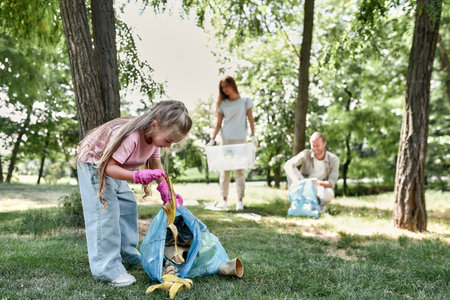 Cute little girl with trash bag collecting garbage while cleaning with parents in the park or forestの写真素材
