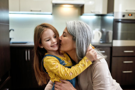 Kisses. Portrait of happy grandmother embracing and kissing her cute little preschool granddaughter, standing together at homeの写真素材