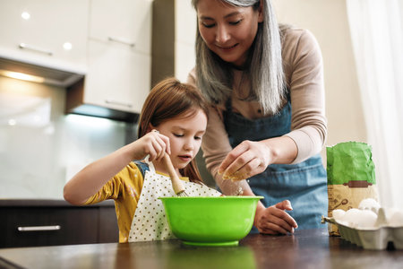 Cute granddaughter whipping with whisk while grandmother adding flourの写真素材