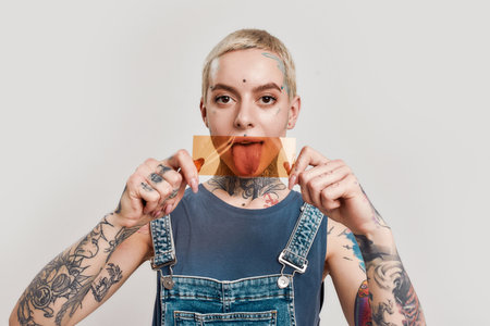 Young girl tattooed with piercing wearing denim overall looking into a camera, holding an orange filter in front of her mouthの写真素材