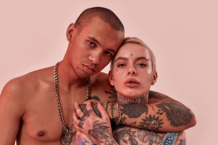 Dark-skinned topless male with tattoos hugging a pierced white tatooed woman, looking into a camera...