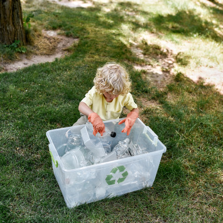 Cute little caucasian boy wearing rubber gloves throwing plastic waste into recycle bin while cleaning in the forest or parkの写真素材