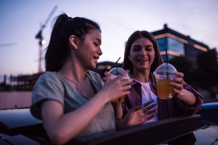 Two girls looking cheerful, holding drinks while sitting in the car and watching a movie in an open air cinema in the eveningの写真素材