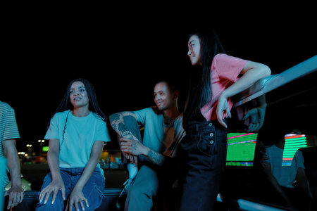 A night meeting of a group of young well-dressed friends of different nationalities sitting in a pick-ups trunk in front of each other outside on a parking site speakingの写真素材