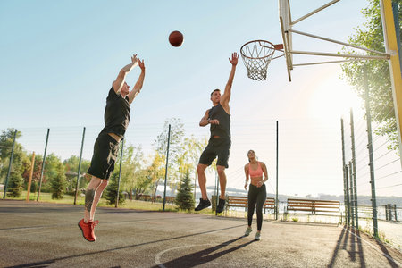 Athletic young friends spending time together outdoors on playground, playing basketball on a sunny...