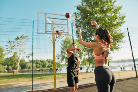 Rear view of sportive young girl throwing ball into basket, while playing basketball with friend outdoors on a sunny dayの写真素材