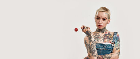 Tattoo and piercing. A white pierced woman with tattoos wearing a denim overall holding a red...