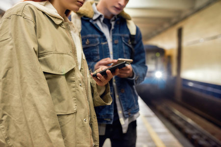 Cropped shot of young people using smartphones while waiting for the train at the subway metro station. Couple of teenagers using mobile app, checking public transport scheduleの写真素材