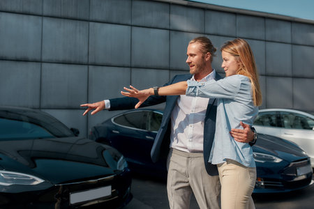 Young couple pointing at car while hugging in showroomの写真素材