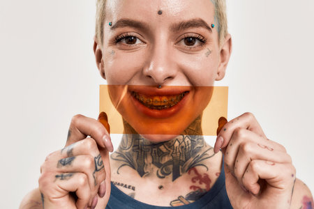 Emotions and postures. A close up of a white girl tattooed and pierced wearing denim overall standing and looking into a camera holding an orange filter in front of her mouth smilingの写真素材