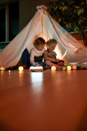 Two little kids, brother and sister reading stories together while sitting on a blanket in a teepee made with bedsheets at homeの写真素材