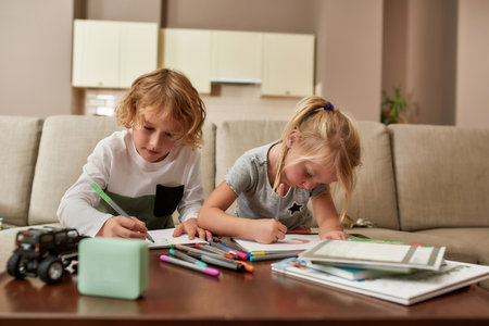 Create. Cute little children, boy and girl looking focused while drawing on paper using marker pen, sitting together on a couch at homeの写真素材
