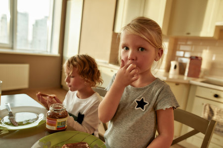 Portrait of adorable little gitl looking at camera, tasting chocolate butter while having breakfast together with her brother in the kitchenの写真素材