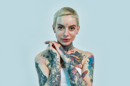 Tattoo and piercing. A tattooed and pierced white girl wearing denim overall standing and looking into a camera holding her hands together near her face and smilingの写真素材