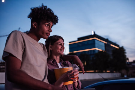 Portrait of attractive young couple, boy and girl spending time together, sitting in the car with popcorn and drink while watching a movie in a drive in cinemaの写真素材