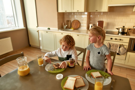 Little children having breakfast on their own in the morning at home. Adorable girl watching her brother making toasted bread with chocolateの写真素材