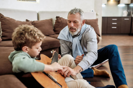 Grandfather teaching his little grandson how to play acoustic guitar while sitting in the living room at homeの写真素材